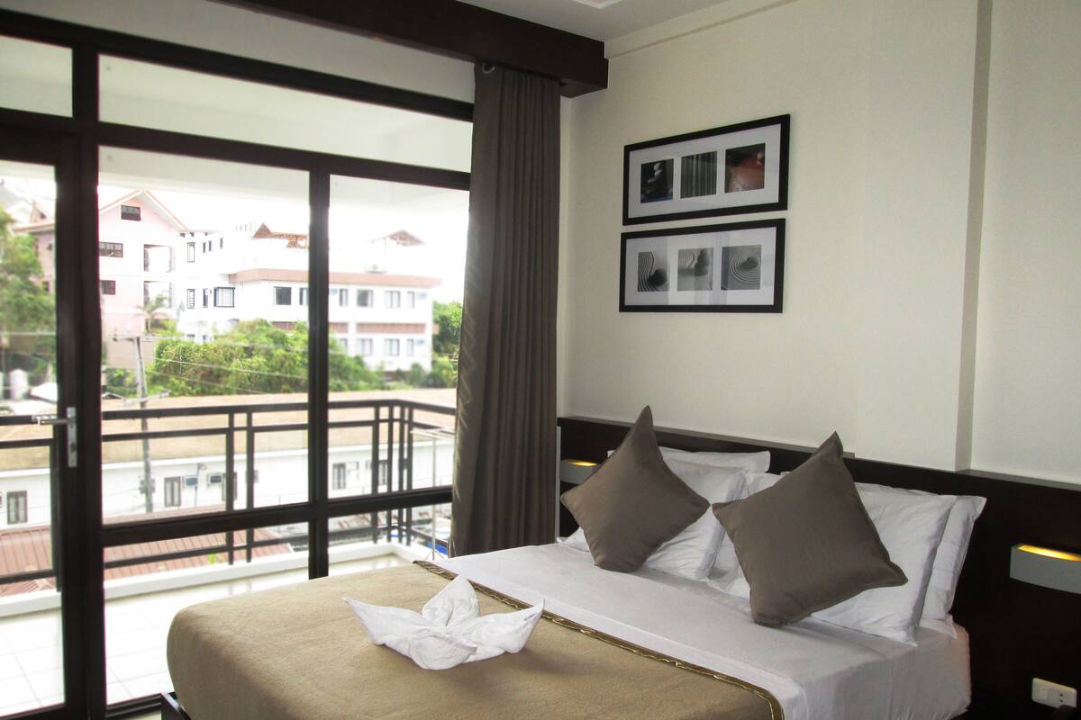 A hotel room with a large window, a bed with white linens and two pillows, a curtain, and two framed pictures on the wall.