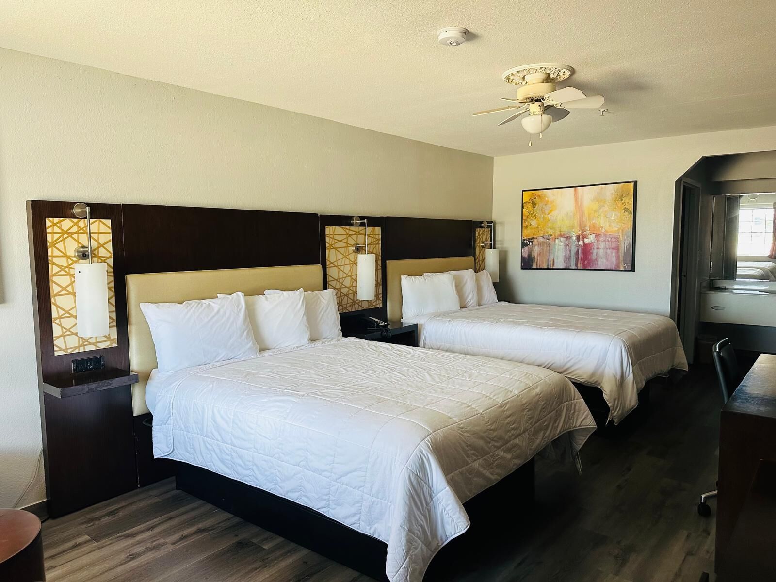 A hotel room with two beds, a ceiling fan, a painting, and a desk.