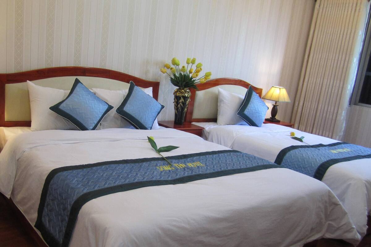A hotel room with two beds, each with white linens and blue quilted bedspreads. There is a wooden headboard, a bedside table with a lamp, and a vase with yellow flowers on the nightstand.