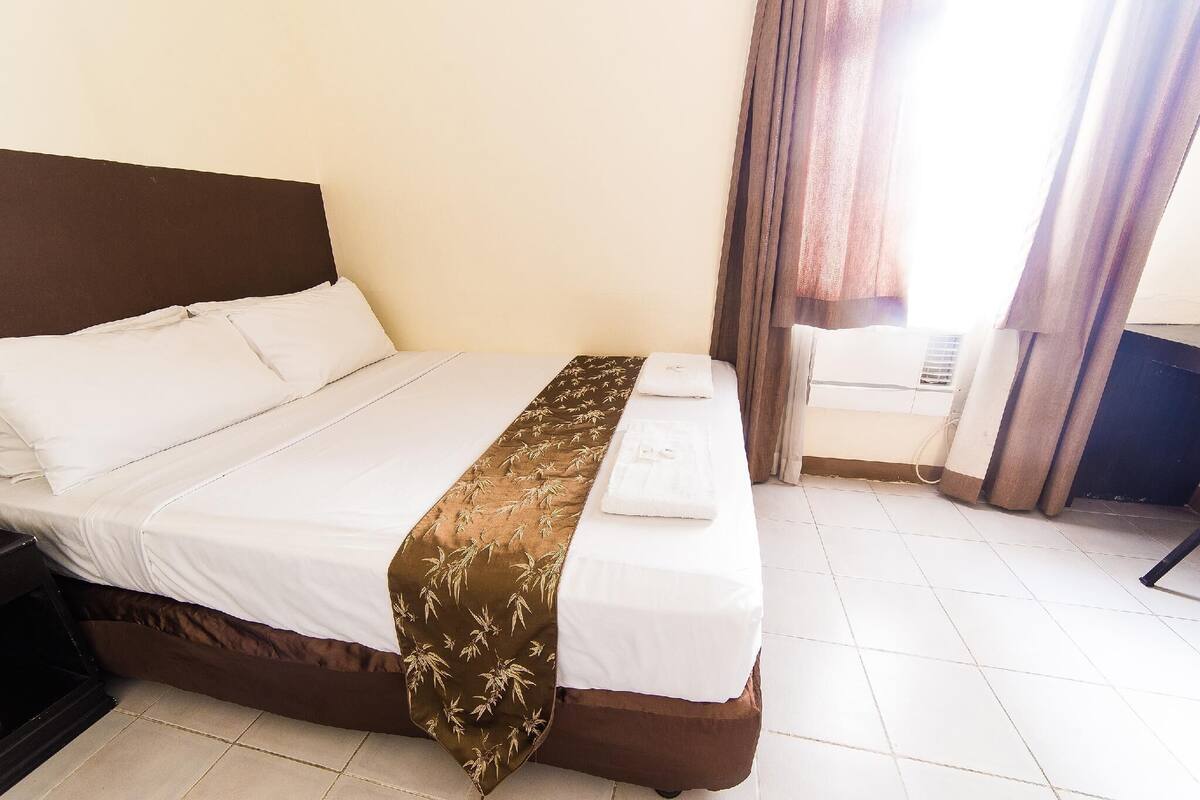A neatly made bed with a brown bedspread and white linens, a bedside table, and a window with curtains.