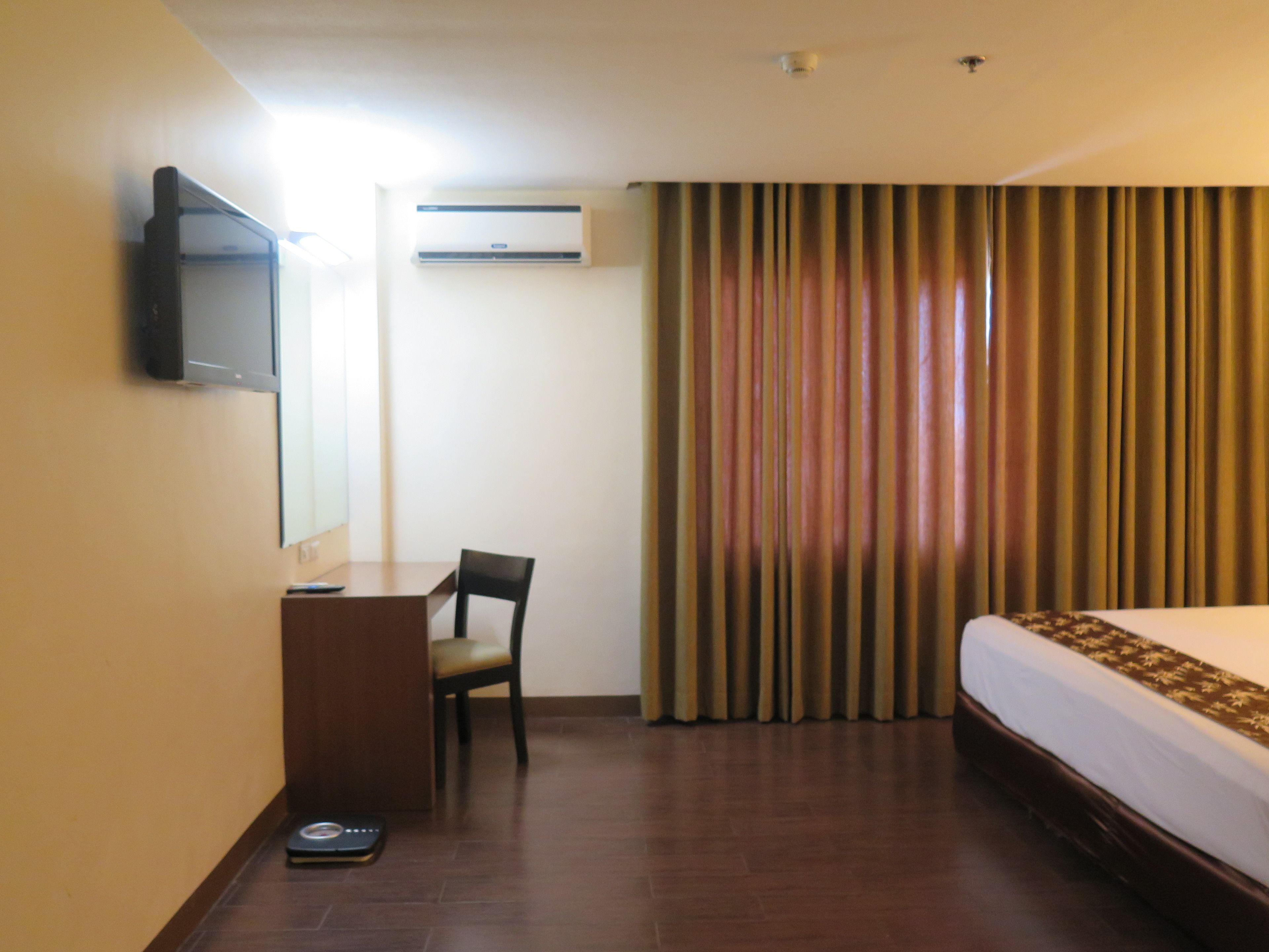 A hotel room with a bed, a desk, a chair, a television, an air conditioning unit, and a window with curtains.