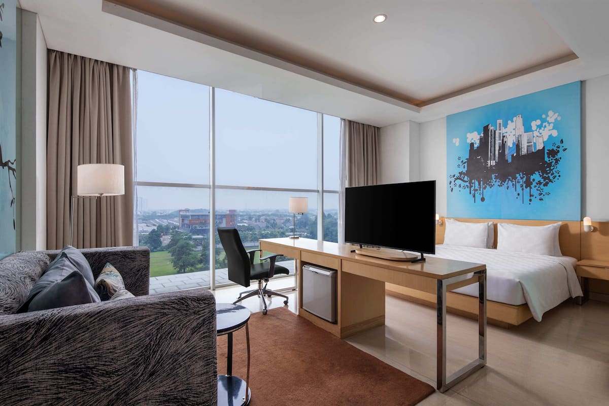 A hotel room with a large window, a desk with a flat-screen TV, a bed, and a painting on the wall.