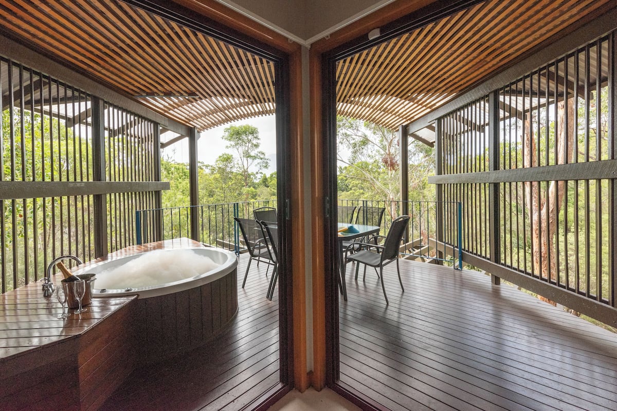 A wooden deck with a hot tub, outdoor seating, and a view of the surrounding greenery.