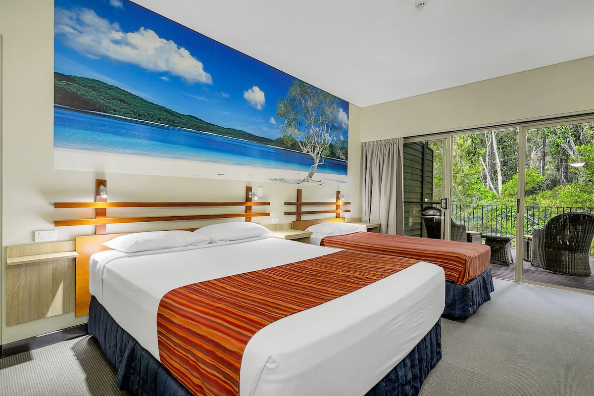 A hotel room with two beds, a large beach mural on the wall, and a balcony with a view of greenery.