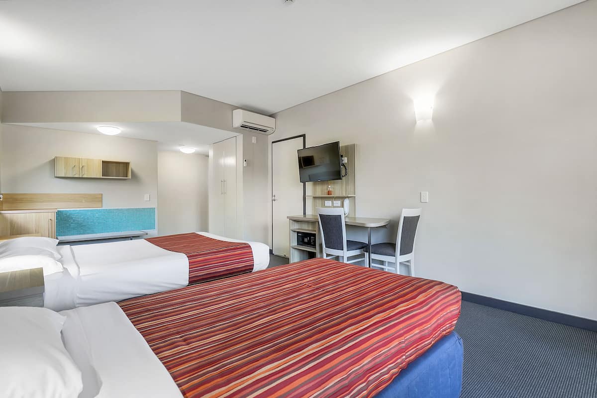 A hotel room with two beds, a desk, a chair, a TV, and an air conditioning unit.