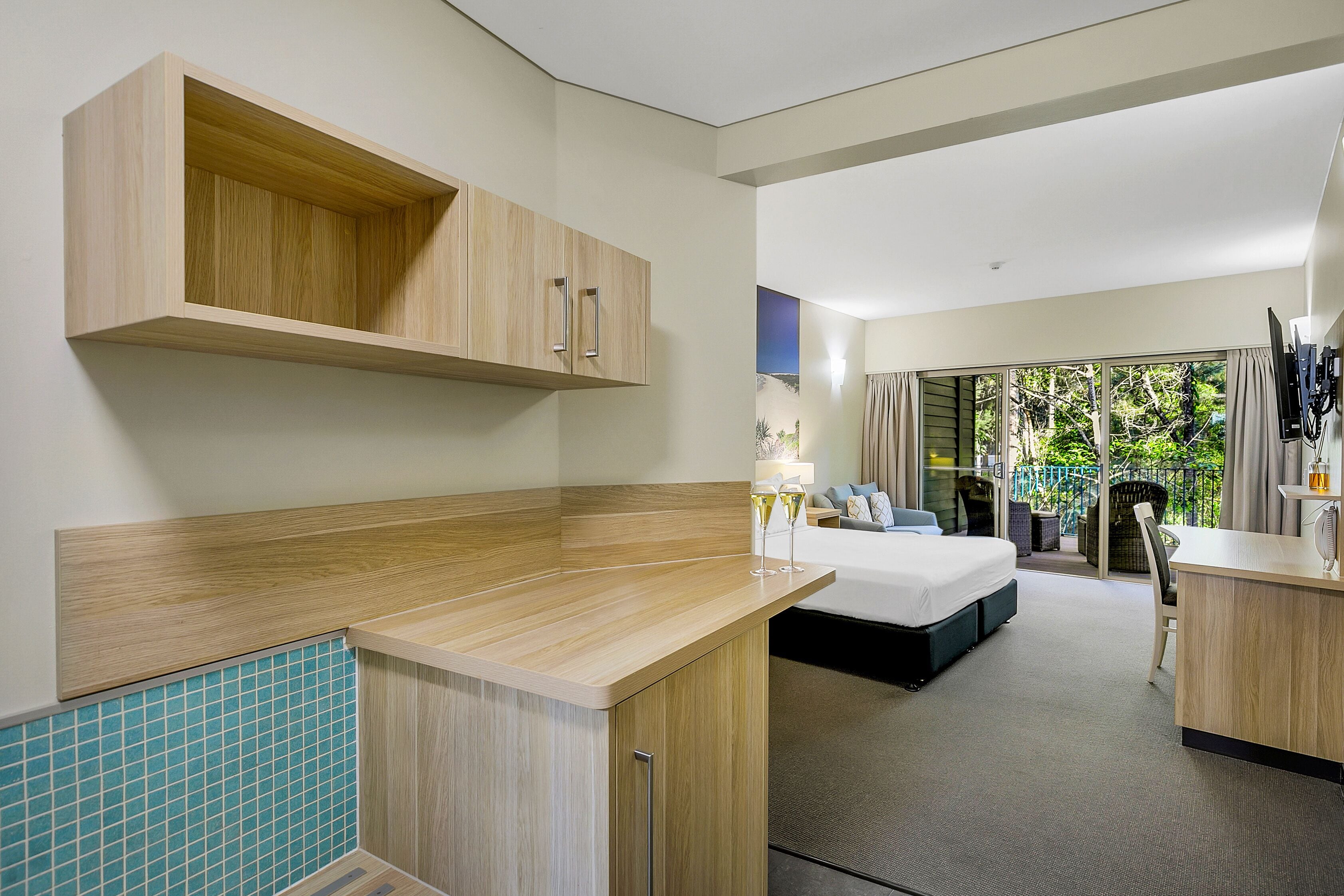 Quarto de hotel moderno com uma kitchenette em madeira, uma cama com vista e uma varanda com mobiliário de exterior.
