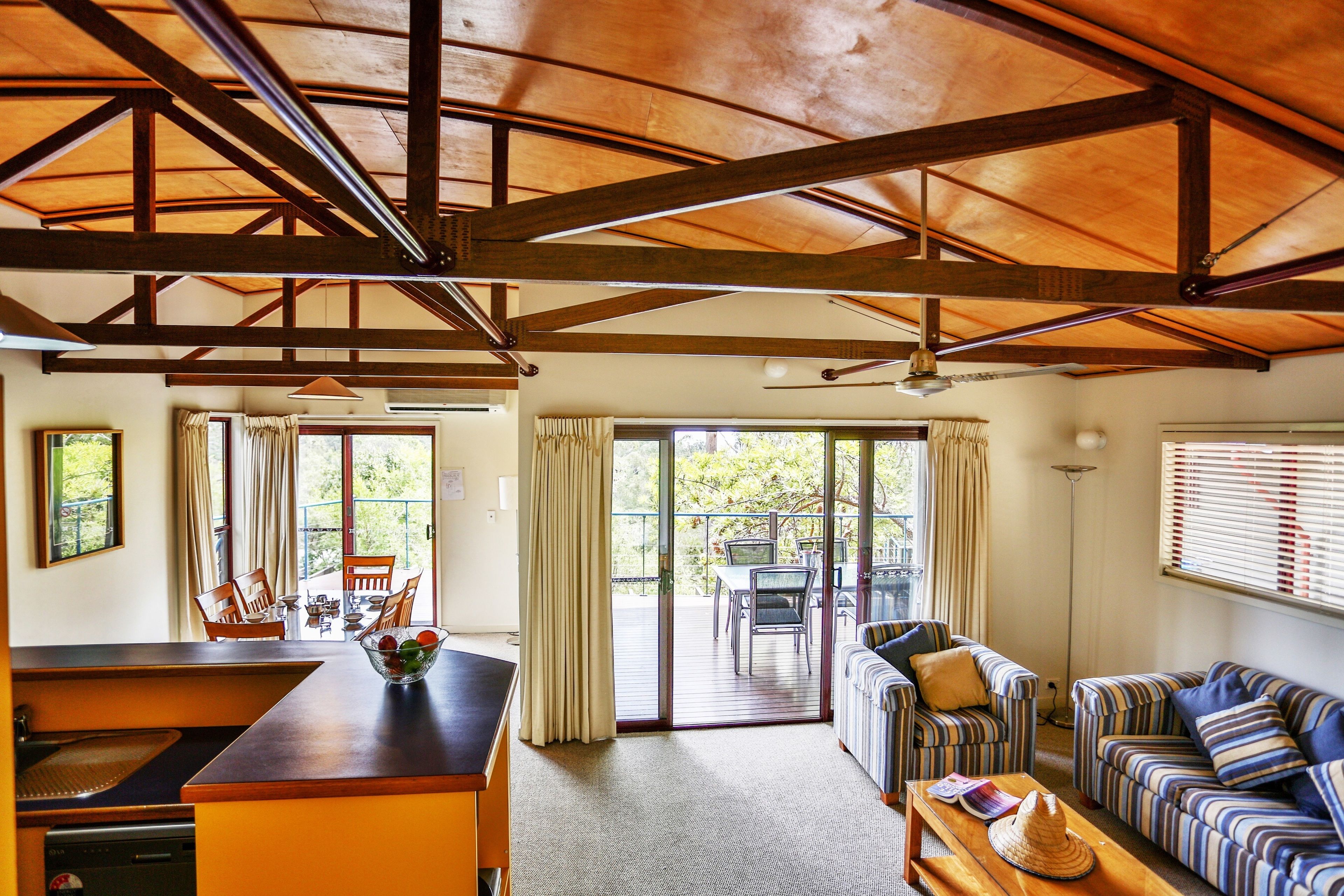 A spacious living area with a wooden ceiling, a dining table with chairs, a sofa set, and a view through the glass doors to the outside.