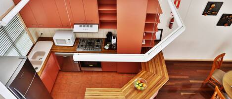 Cocina compacta con microondas, estufa y pileta. Hay un banco de madera con un frutero y un comedor con sillas.
