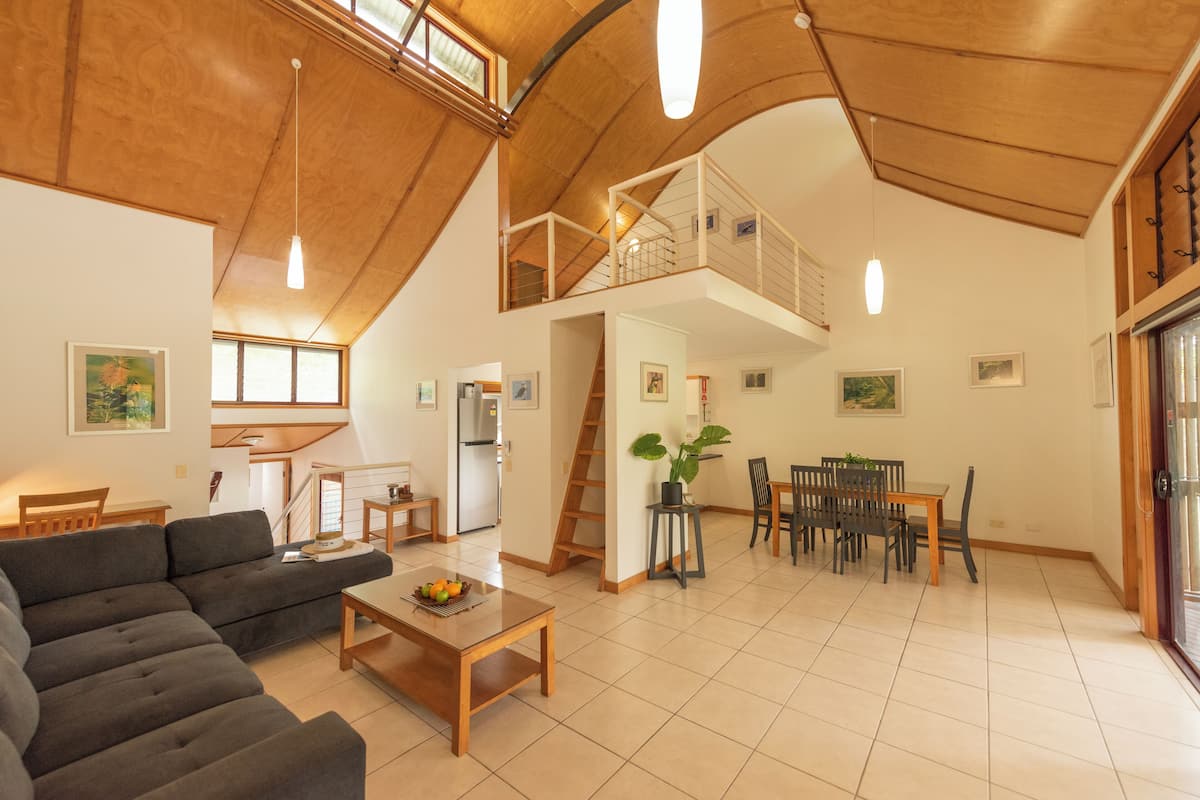 A spacious living area with a high ceiling, a dining table, a sofa, and a staircase leading to an upper level.