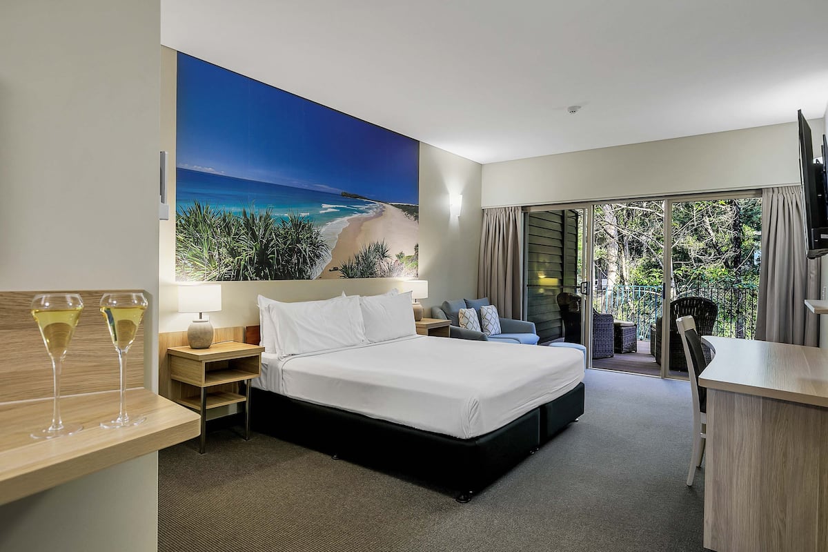 A hotel room with a bed, a desk, a chair, a sofa, a TV, and a balcony with a view of the beach.