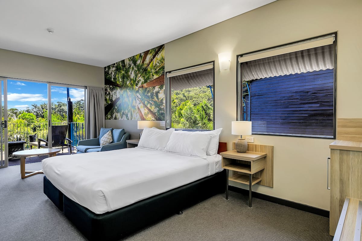 A modern hotel room with a large bed, a seating area with a table and chairs, a desk with a lamp, and large windows offering a view of greenery.