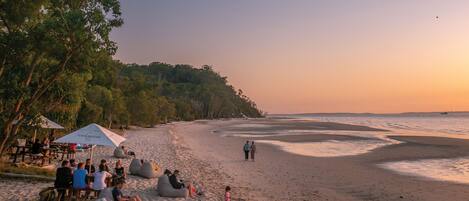 Ein Strand mit Personen, die auf Sitzsäcken sitzen, und einigen Spaziergängern. Sonnenschirme und ein Tisch mit Personen darum.