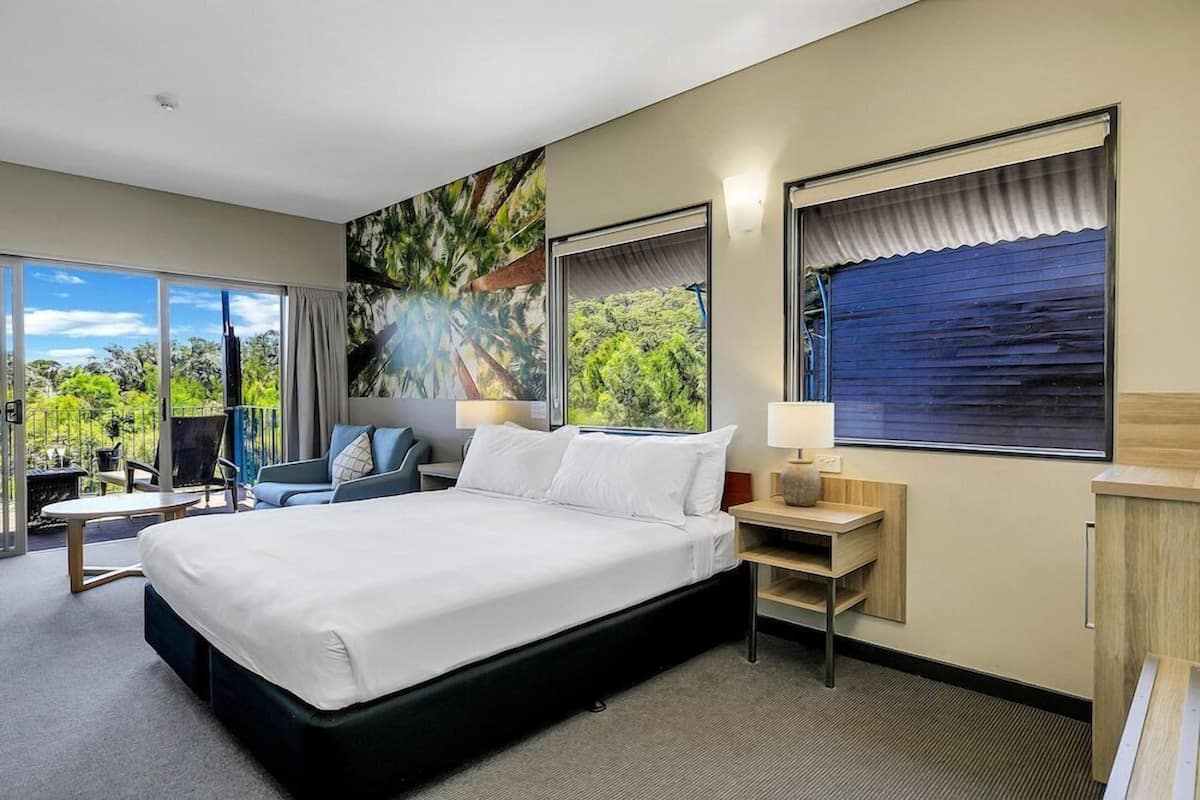 A hotel room with a bed, a desk, a chair, a small table, and a large window with curtains.