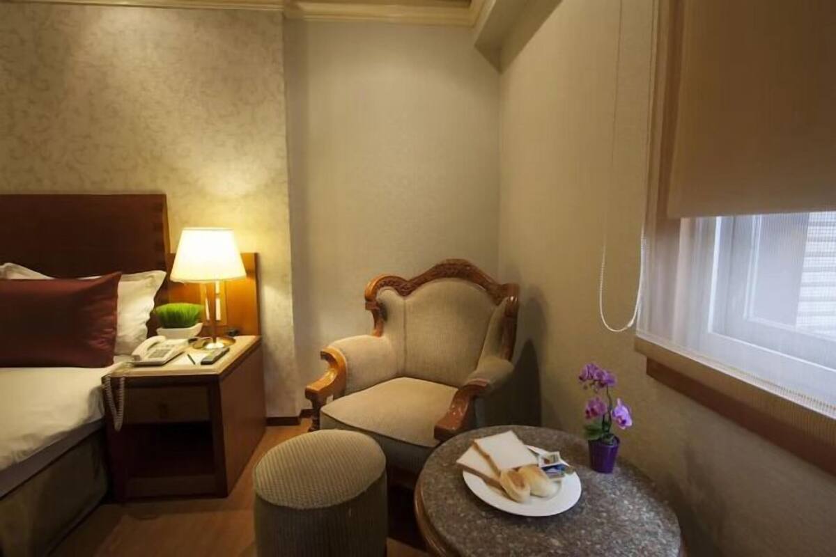 A hotel room with a bed, a chair, a small table with a plate and a vase of flowers, a nightstand with a lamp, and a window with blinds.