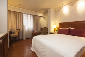 Une chambre d’hôtel avec un grand lit, une tête de lit en bois, un bureau avec une chaise, une petite table avec une lampe et une fenêtre avec des rideaux.