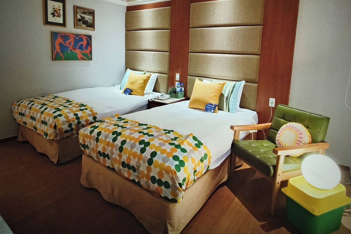 Ein Hotelzimmer mit zwei Betten, einem grünen Sessel, einem kleinen Tisch und einer Wand mit Kunstwerken.