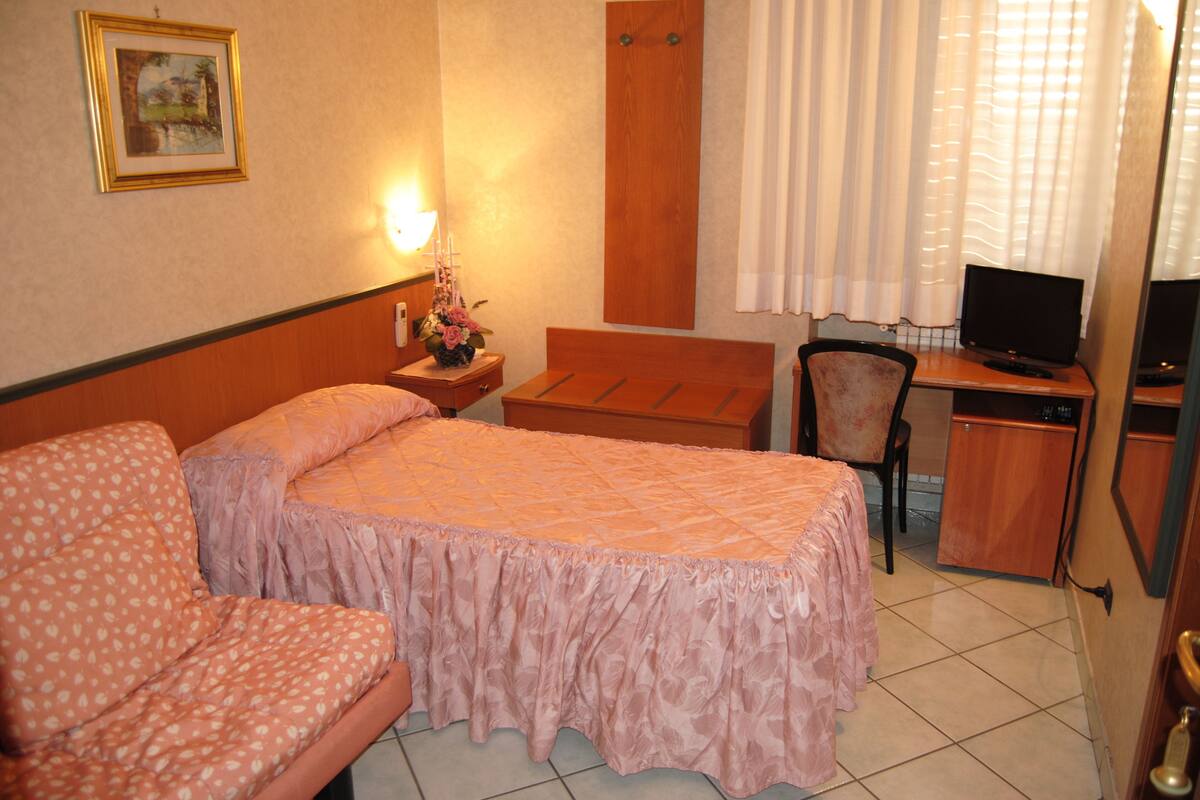 A hotel room with a bed, a chair, a desk with a computer, a lamp, and a painting on the wall.