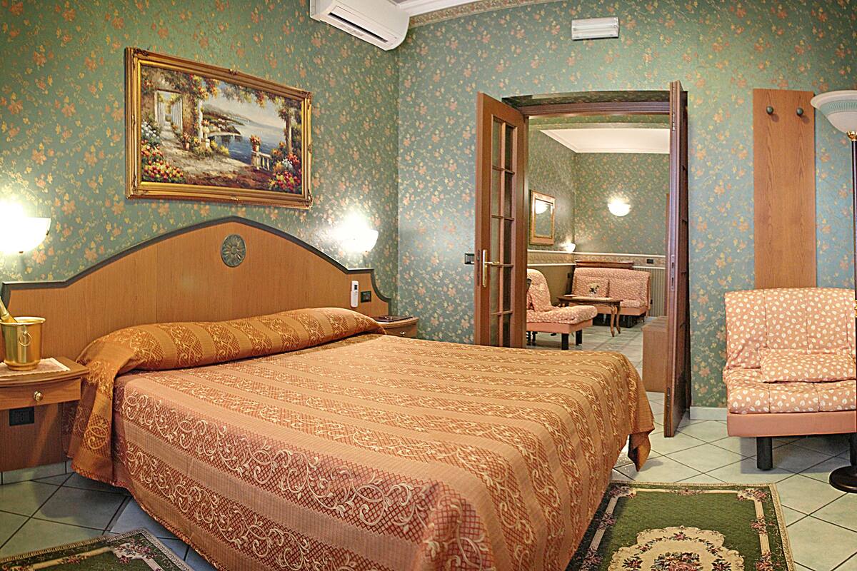 A hotel room with a large bed, a nightstand, a lamp, a chair, and a painting on the wall.
