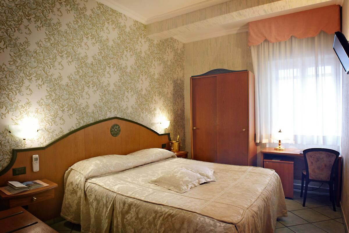 A hotel room with a bed, a desk, a chair, a wardrobe, and a nightstand with a lamp.