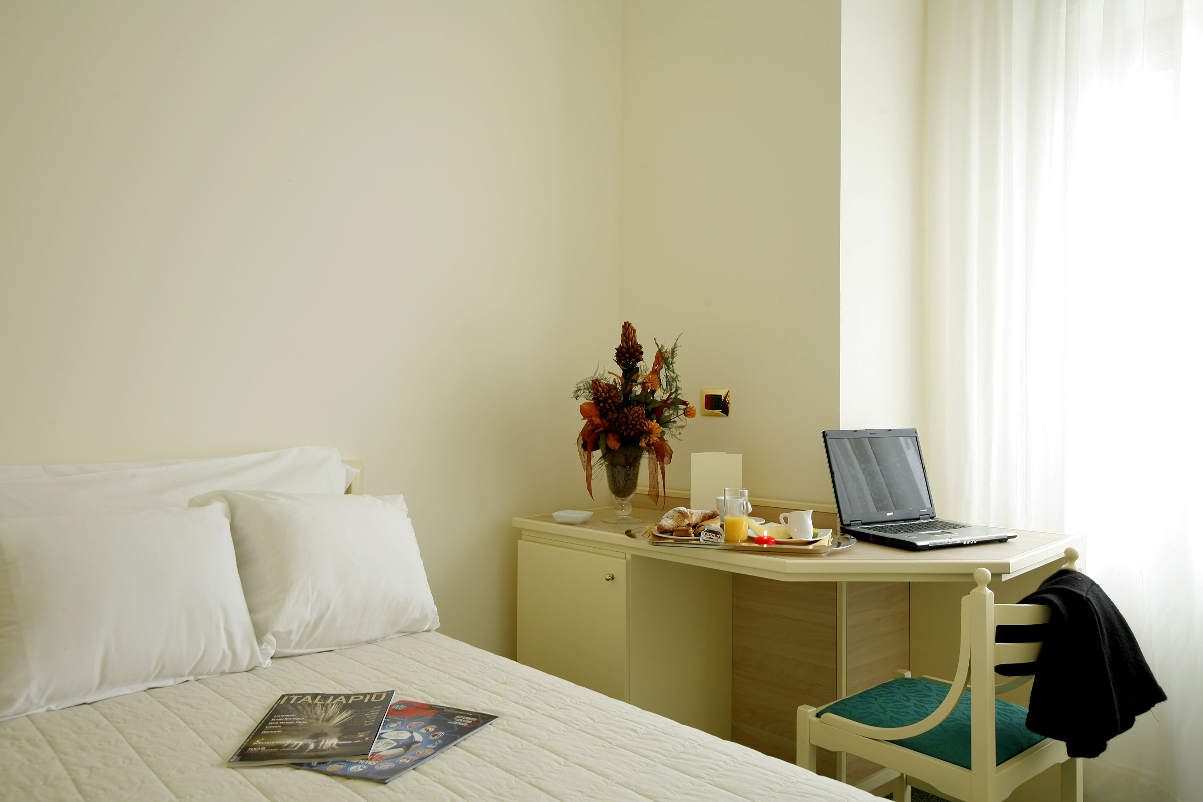 Habitación de hotel con una cama, un escritorio con una computadora portátil, una silla, un florero con flores y una revista sobre la cama.