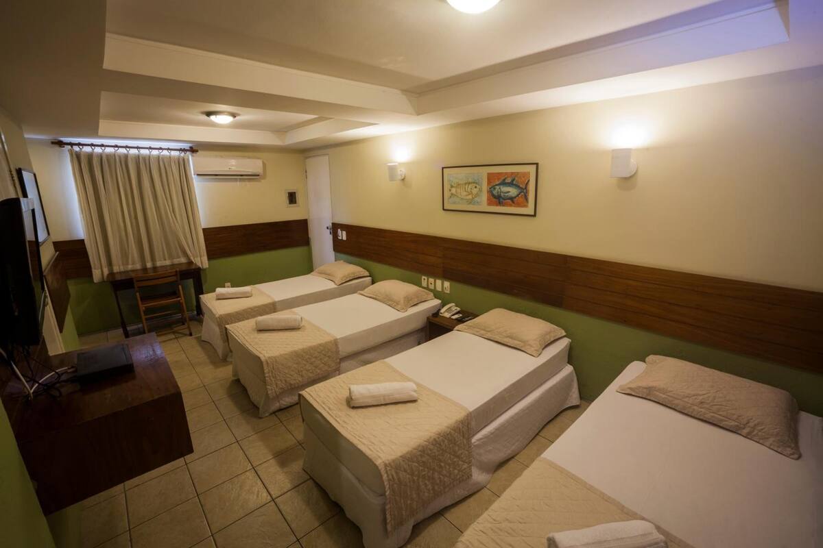 Quarto de hotel com três camas de solteiro, uma escrivaninha com computador, uma cadeira, televisão e aparelho de ar condicionado.