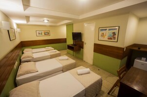 Quarto de hotel com quatro camas de solteiro, televisão, pequena mesa de trabalho e obras de arte nas paredes.