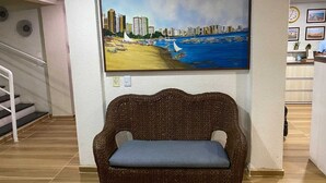 A living room with a large painting of a coastal cityscape, a wicker sofa with a cushion, a staircase, and a reception area with framed pictures on the wall. - Hotel Casa de Praia (Fortaleza)