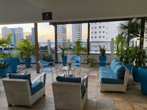 Un área de estar exterior cubierta con mobiliario de mimbre y plantas en macetas, con vistas a un paisaje urbano.