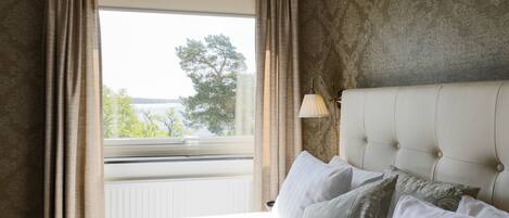 A hotel room with a large bed, a window with curtains, a lamp, and a view of greenery outside.