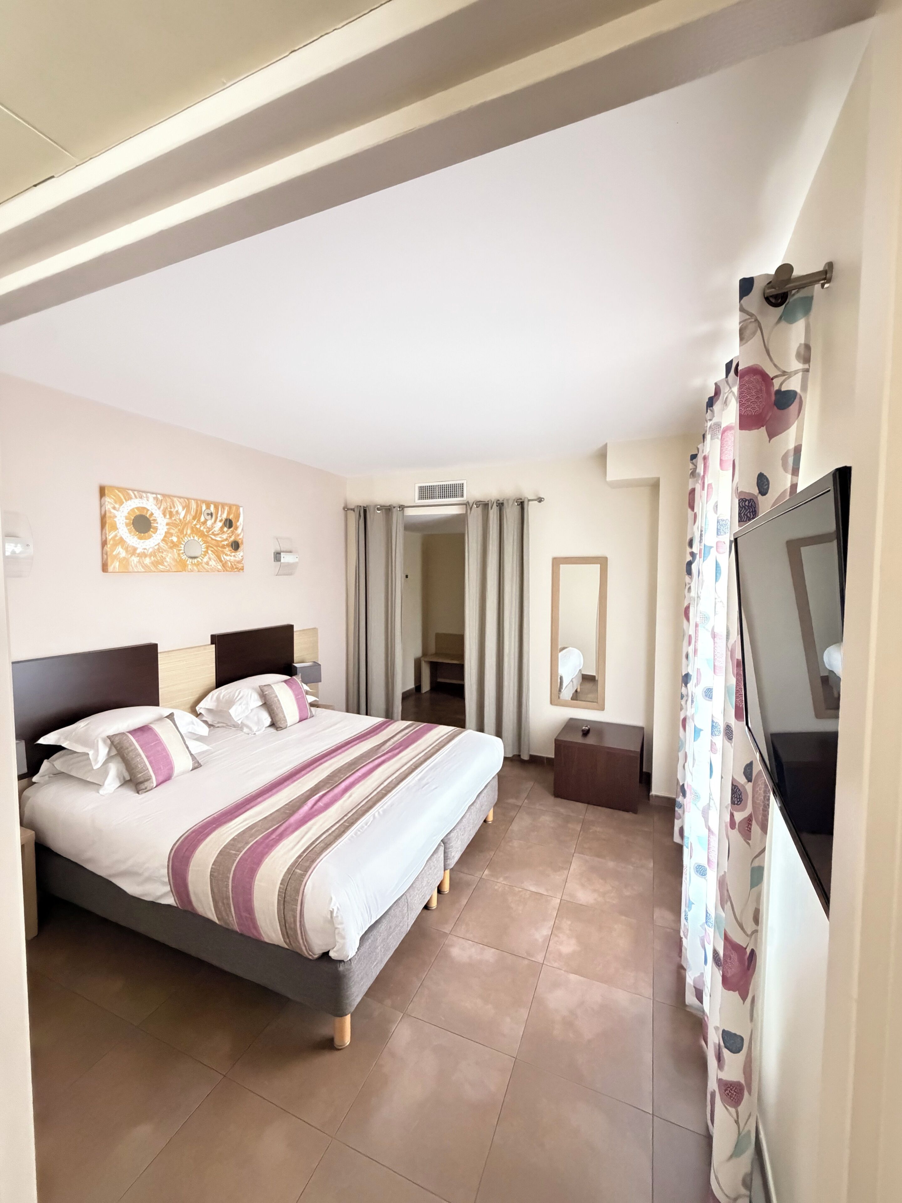A hotel room with a bed, a TV, a mirror, and a door leading to another room.