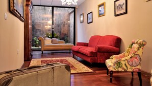 Ein Wohnzimmer mit einem roten Sofa, einem gemusterten Sessel und einem beigen Sofa. Es gibt eine Chandelüster, gerahmte Bilder an der Wand und einen Ausblick durch eine Glastür.