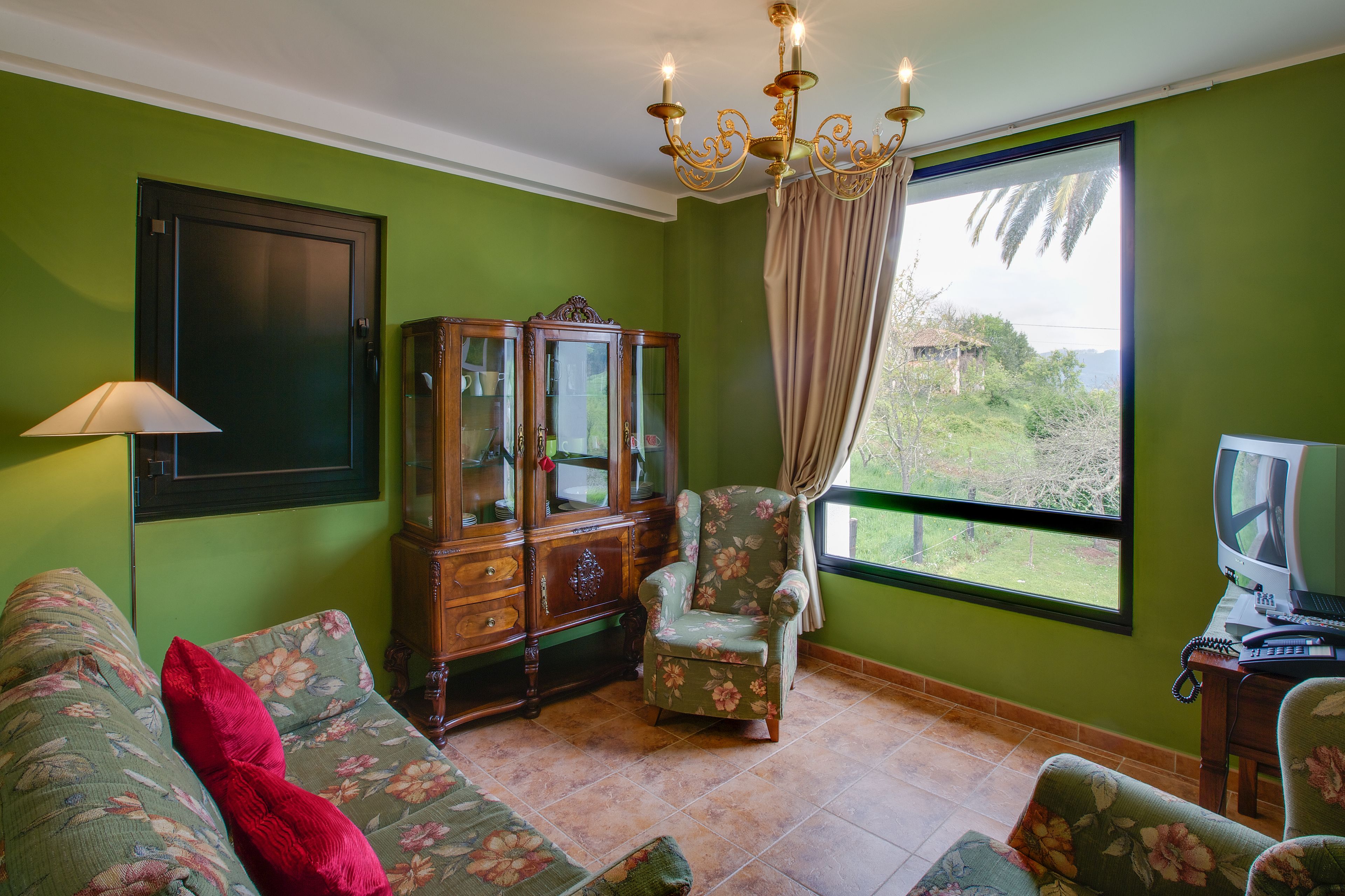 A room with a green wall, a wooden cabinet, a floral armchair, a sofa with a red pillow, and a television on a wooden table.