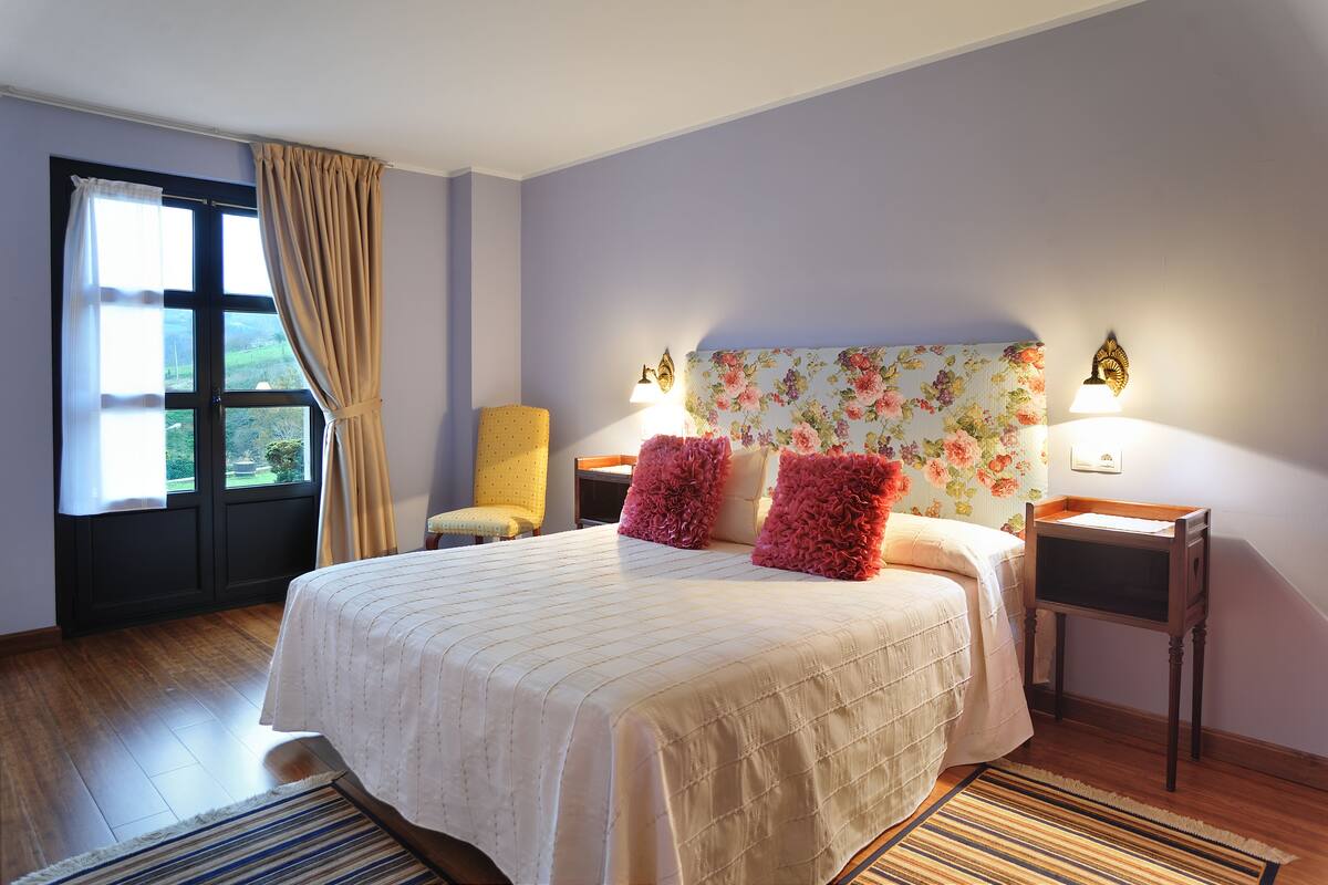 Une chambre à coucher avec un grand lit, deux tables de chevet, une chaise, une fenêtre avec des rideaux et une tête de lit à motifs floraux.