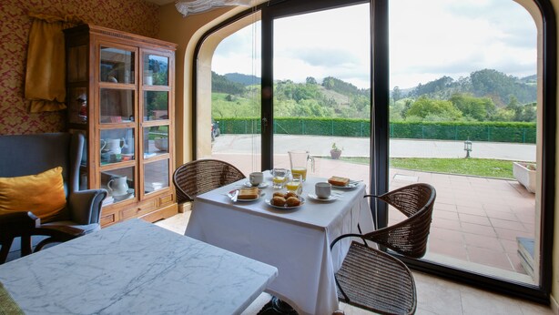 Ein Essbereich mit einem für das Frühstück gedeckten Tisch, einer Vitrine mit Glastüren, Korbstühlen und Blick auf eine grüne Landschaft durch große Fenster.