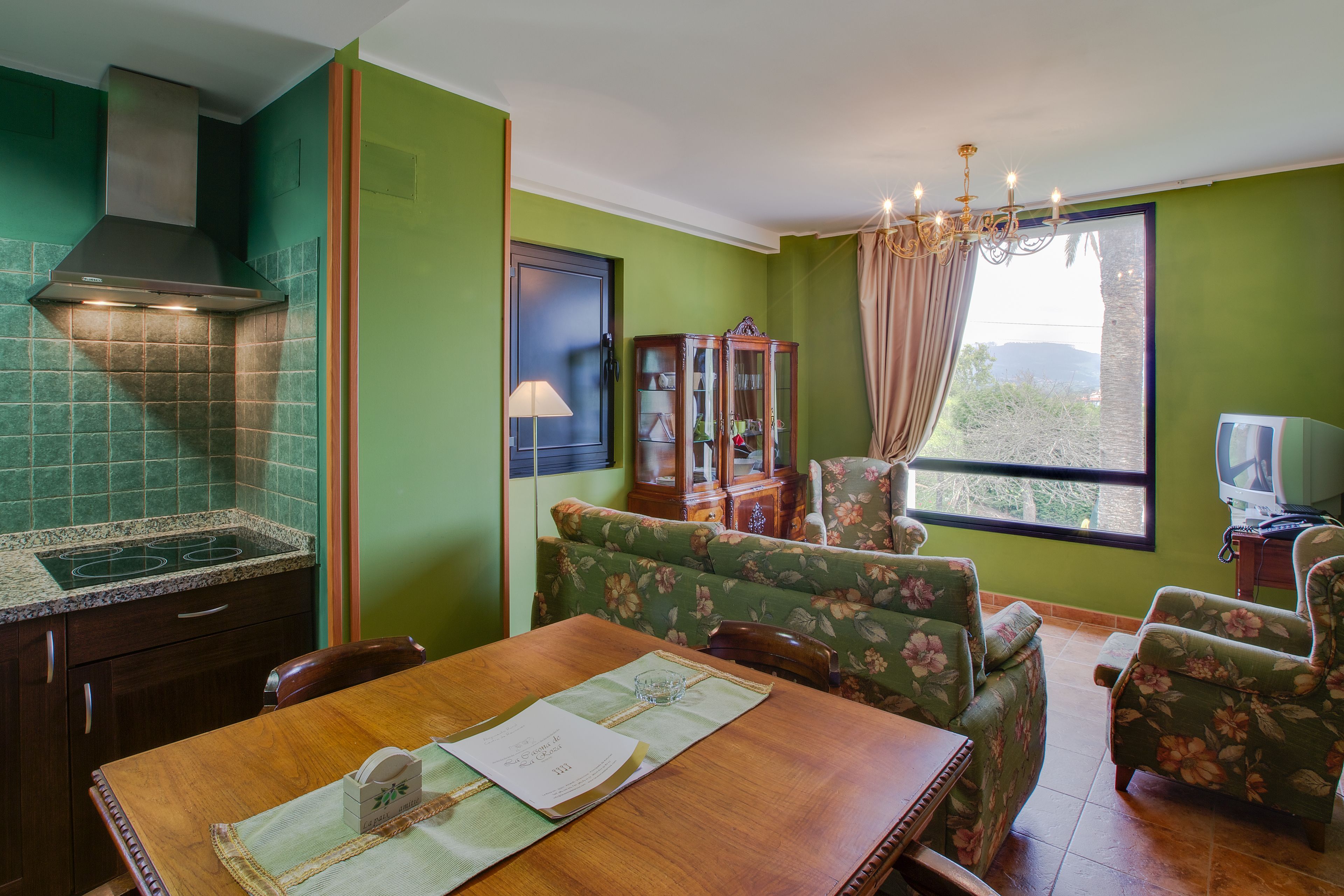Un comedor con una mesa de madera, sillas tapizadas con motivos florales, una araña y vistas a través de una ventana.