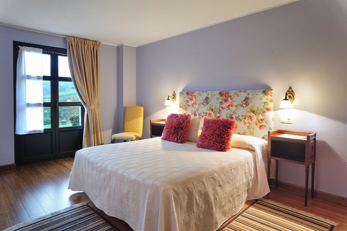 Een slaapkamer met een groot bed, twee nachtkastjes, een stoel, een raam met gordijnen en een hoofdbord met bloemenmotief.