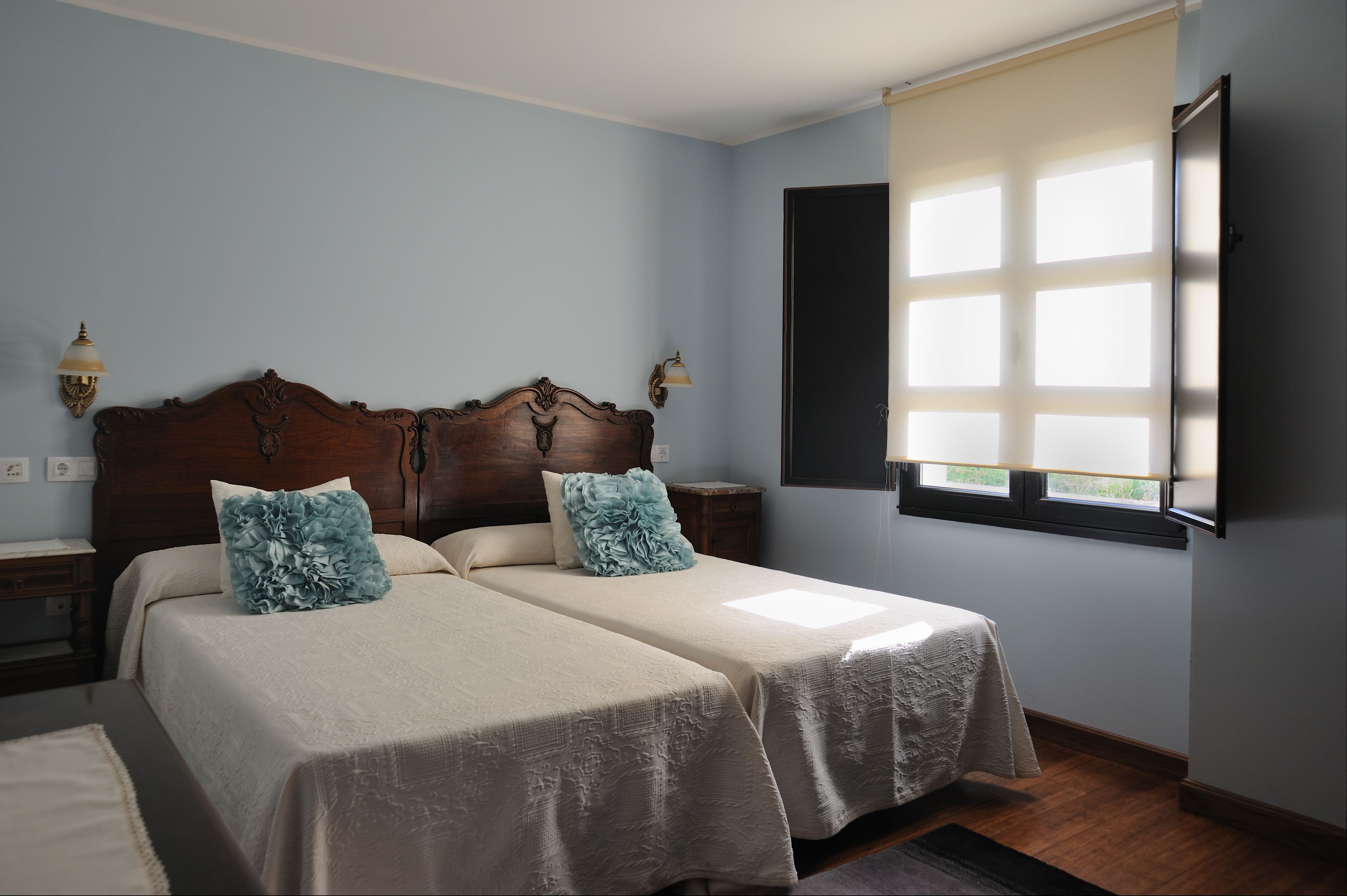 A bedroom with two beds, a wooden headboard, bedside lamps, a window with blinds, and a nightstand.