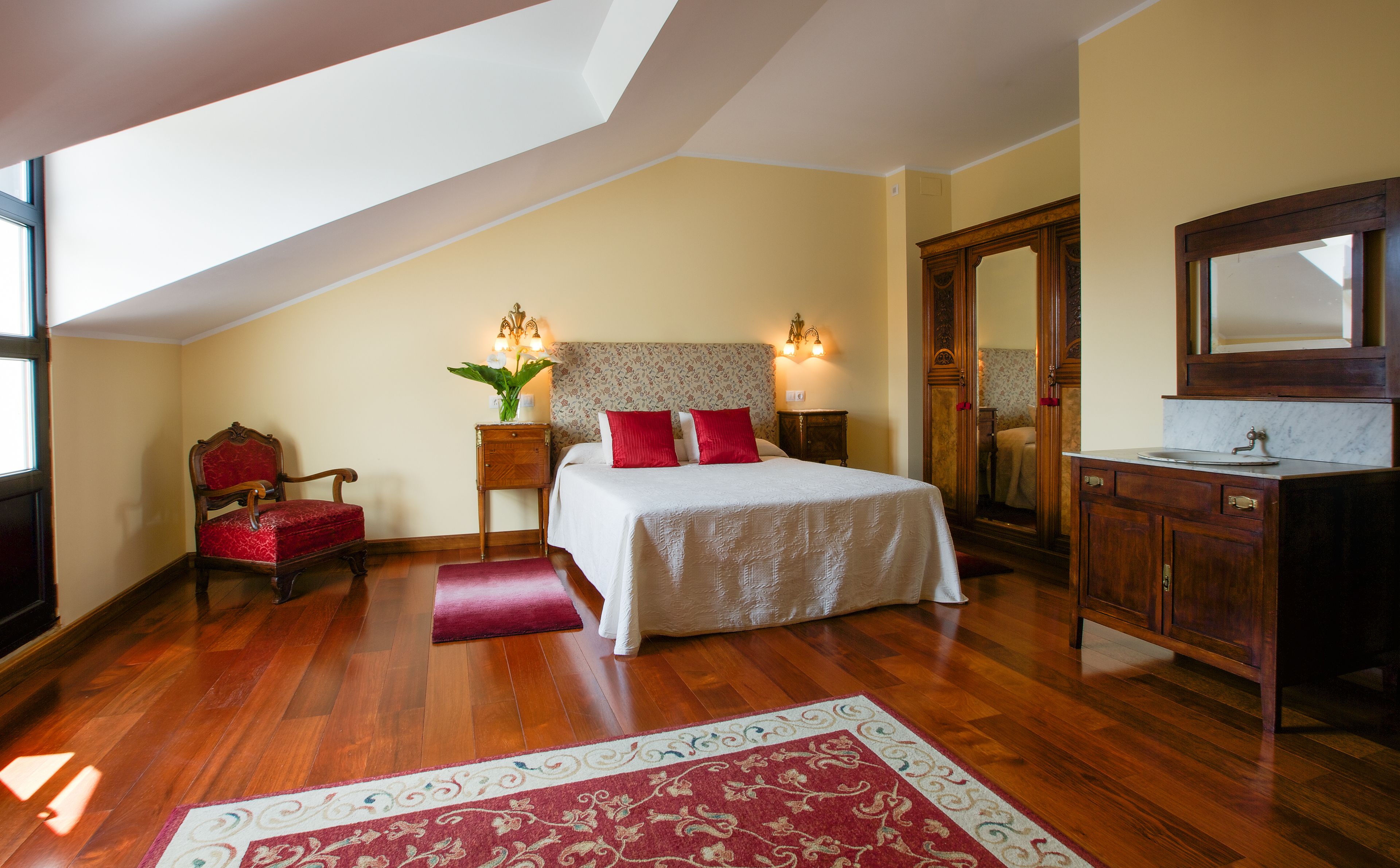 A bedroom with a sloped ceiling, a bed with red and white bedding, a wooden chair, a nightstand with a lamp, a mirror, and a wooden dresser with a sink.
