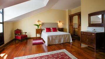 A bedroom with a sloped ceiling, a bed with red and white bedding, a wooden chair, a nightstand with a lamp, a mirror, and a wooden dresser with a sink.