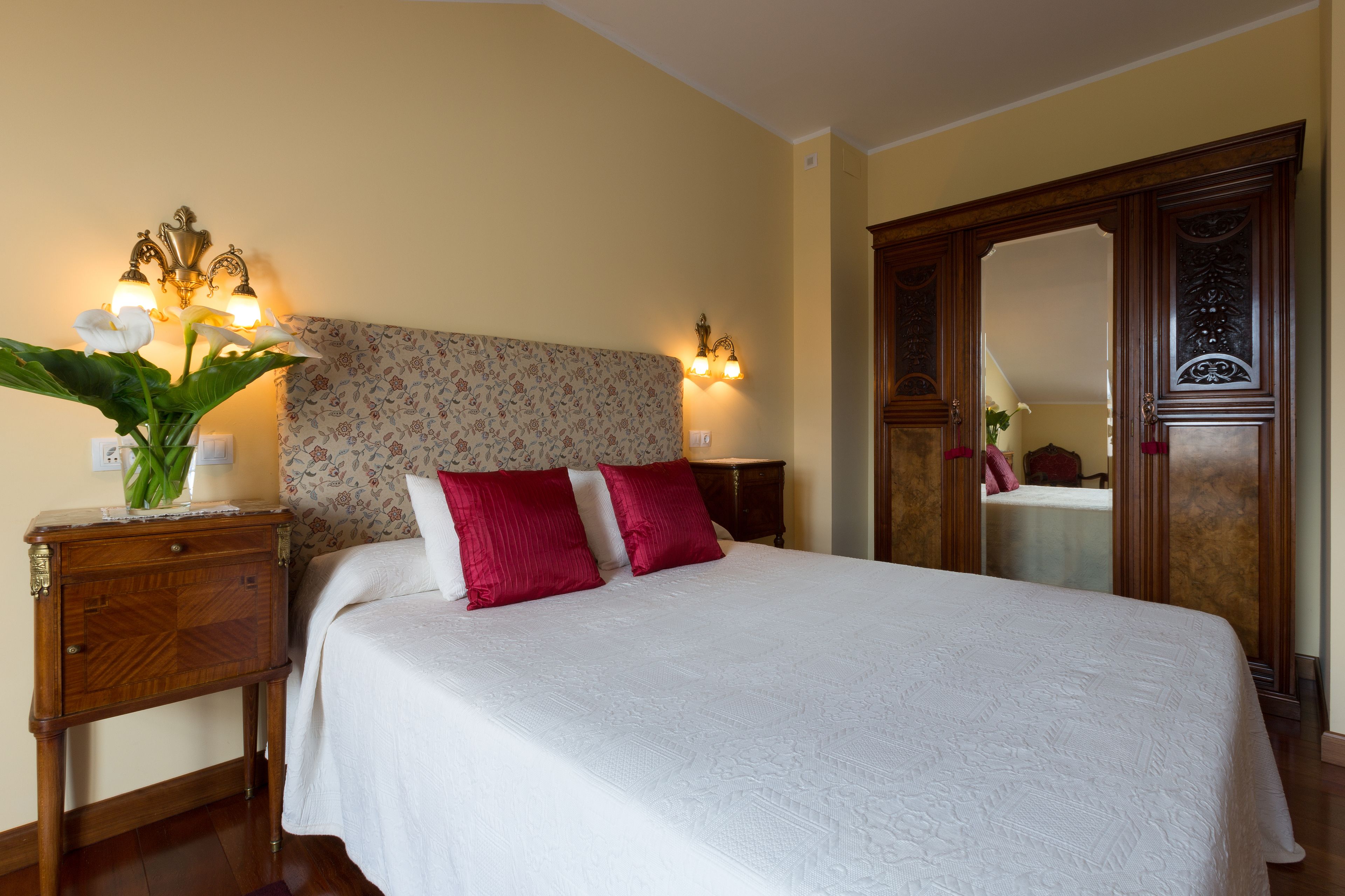 A neatly made bed with white and red pillows, a floral headboard, and a bedside table with a vase of flowers.