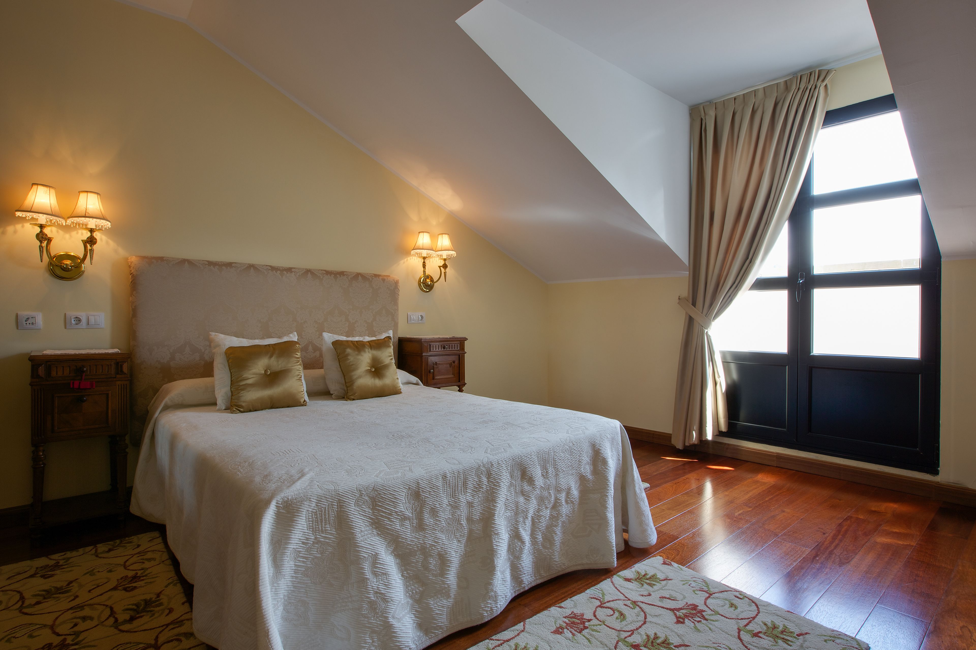 A bedroom with a large bed, two bedside tables, wall sconces, a window with curtains, and a wooden floor.