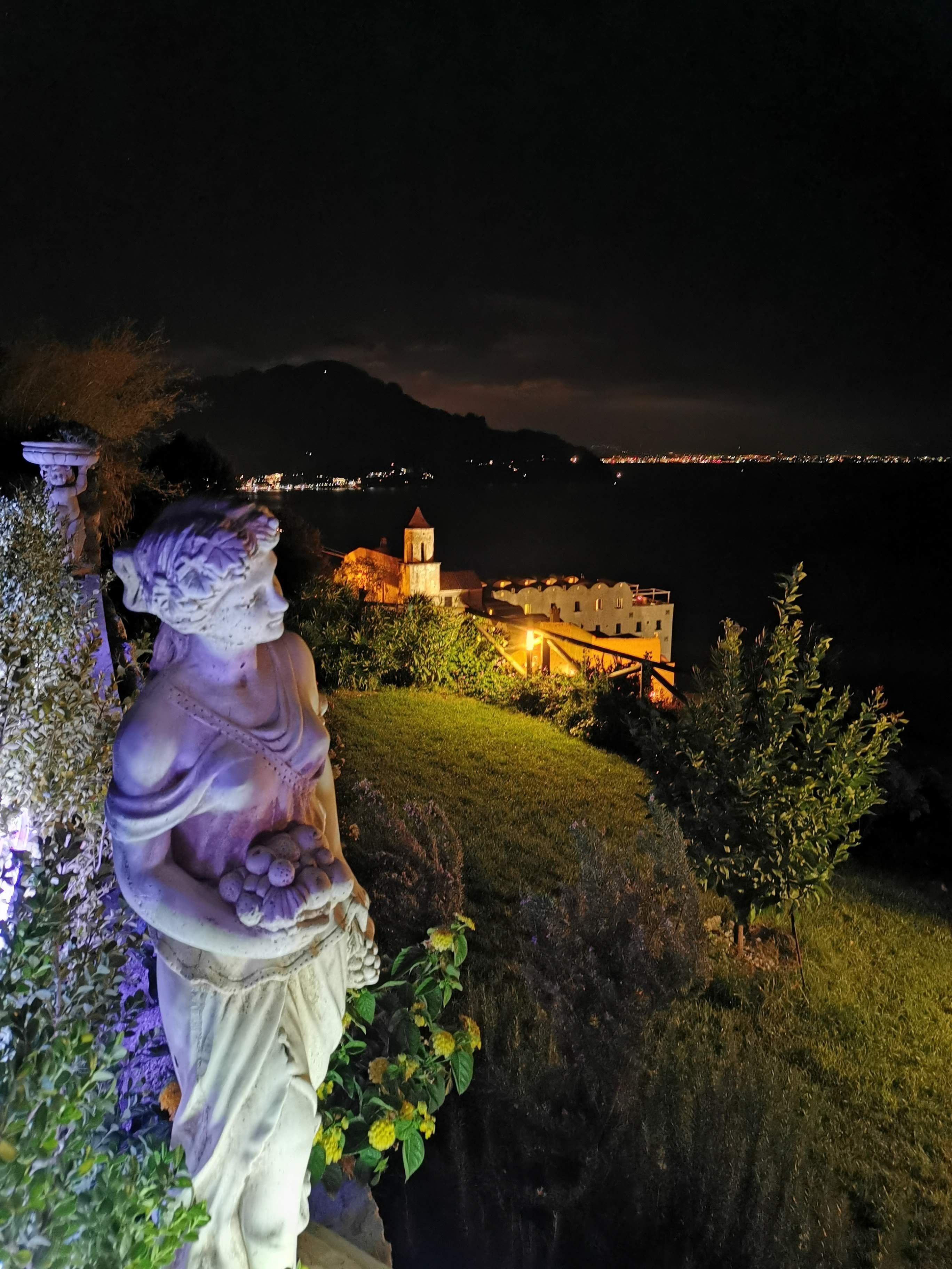 Estátua em um jardim, com vista para um edifício histórico e um corpo de água à noite.