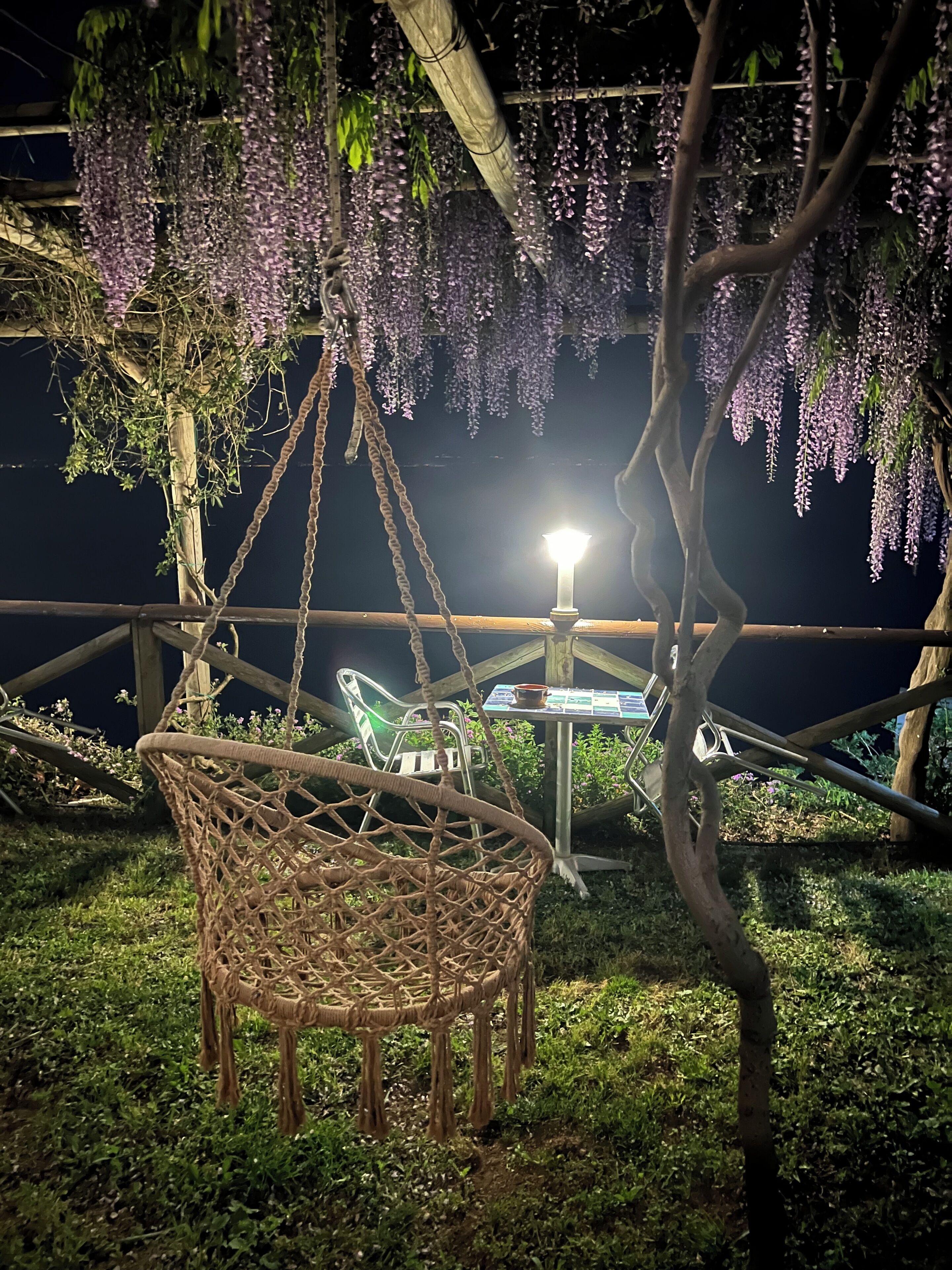Une chaise en osier placée sous une sorte de dais floral suspendu, de couleur violette, une table avec une bougie et une chaise éclairée par une lumière néon.