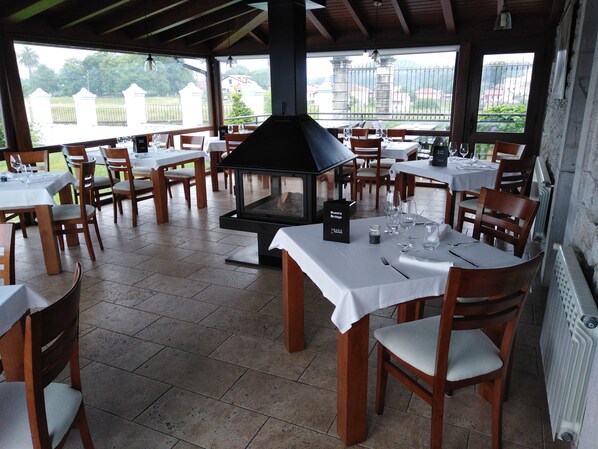 An outdoor dining area with tables set for two, a wood-burning stove, and a view of the surrounding landscape. - Hotel Villadesella (Ribadesella)
