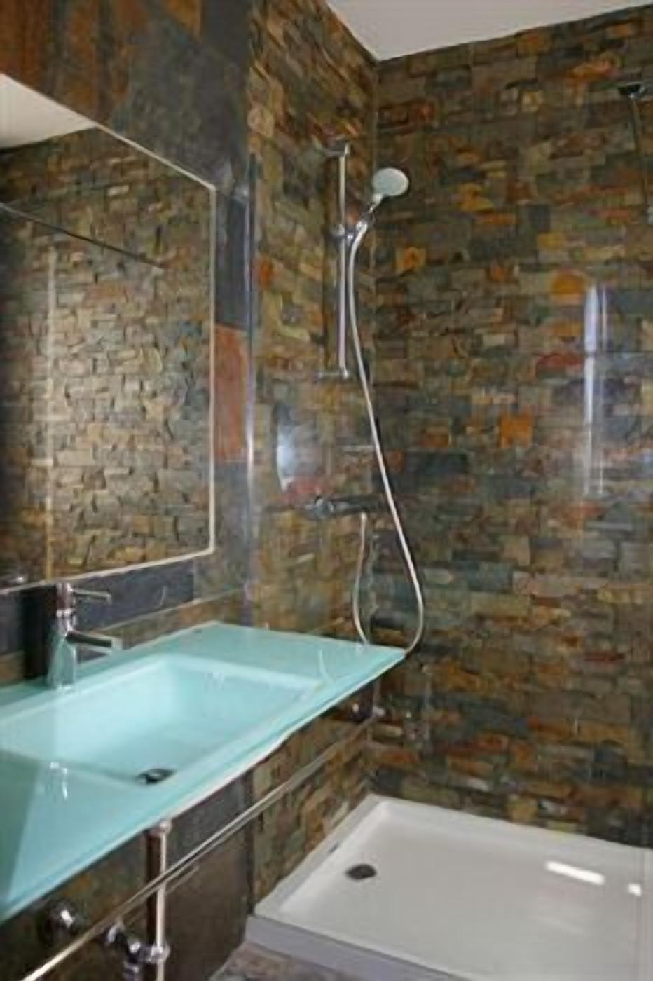 A bathroom with a stone mosaic wall, a glass sink, and a shower.