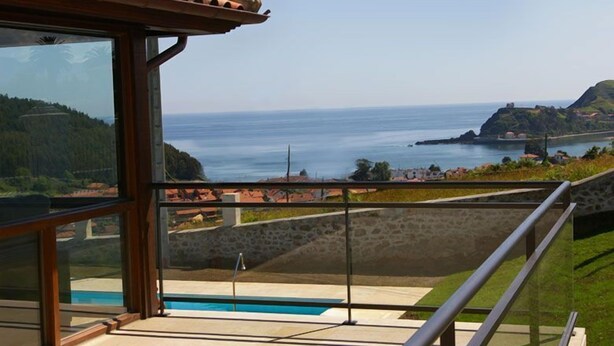 A balcony with a glass railing overlooking a coastal view, a swimming pool, and a stone wall. - Hotel Villadesella (Ribadesella)