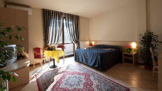 A bedroom with a bed, a desk, a chair, a table with flowers, a window with curtains, and a potted plant. - Villa Giovanna (Somma Lombardo)