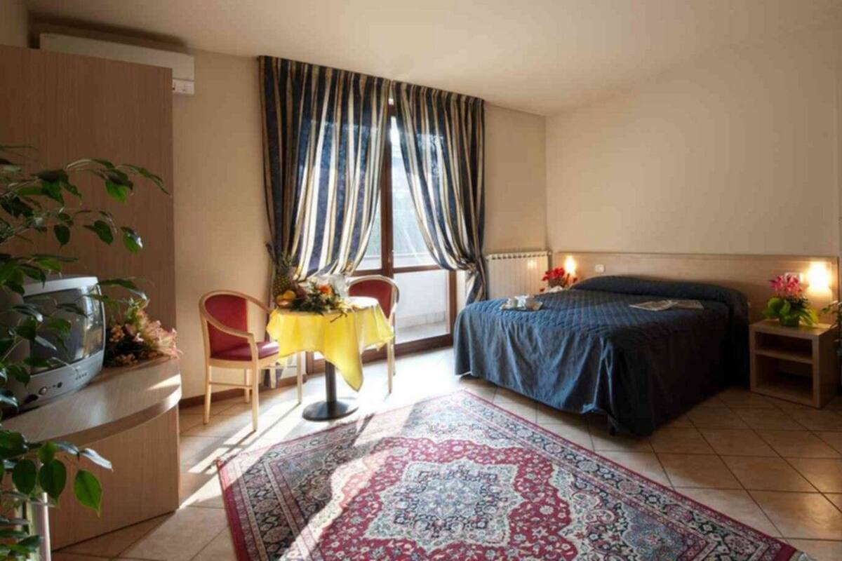 A bedroom with a bed, a desk, a chair, a table with flowers, a window with curtains, and a potted plant.