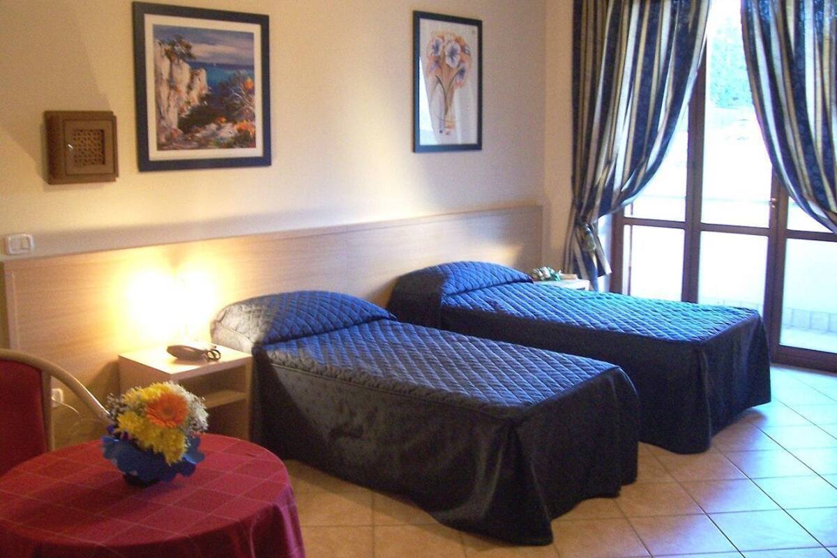 A hotel room with two beds, a small table with a vase of flowers, a nightstand, and a window with curtains.