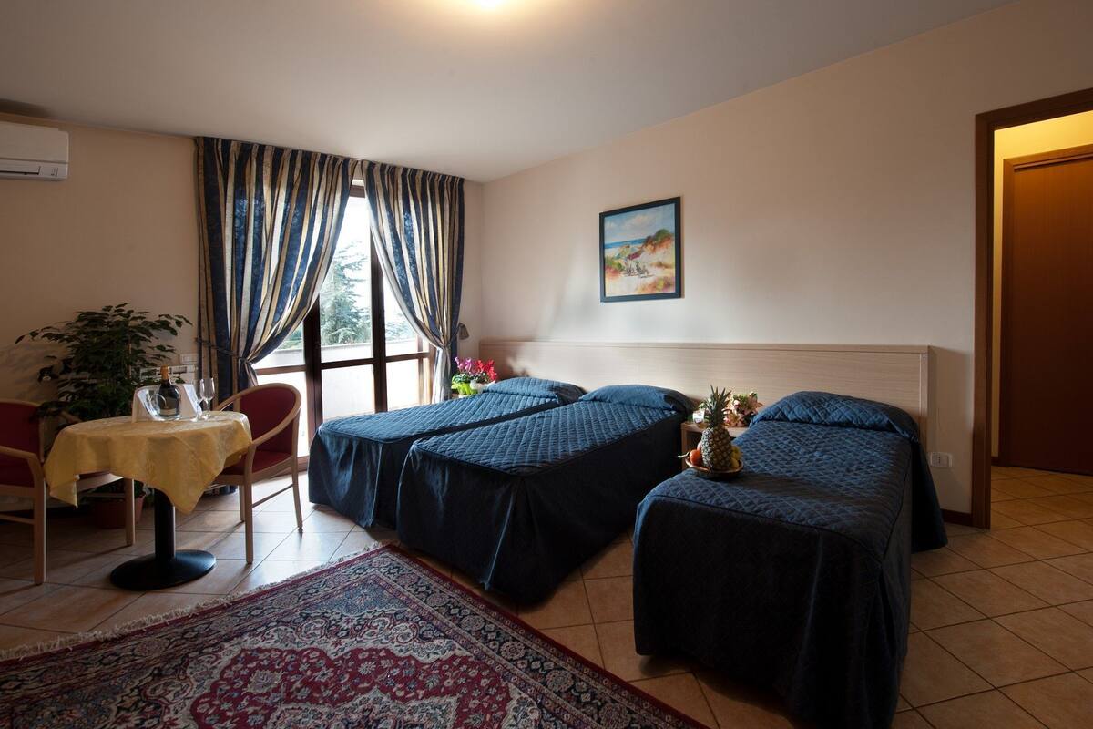 A hotel room with two beds, a dining table, a chair, a large window with curtains, a painting on the wall, and a rug.